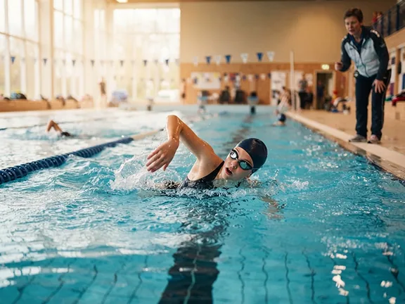 Jonge zwemster in borstcrawl terwijl de coach vanaf de badrand meekijkt